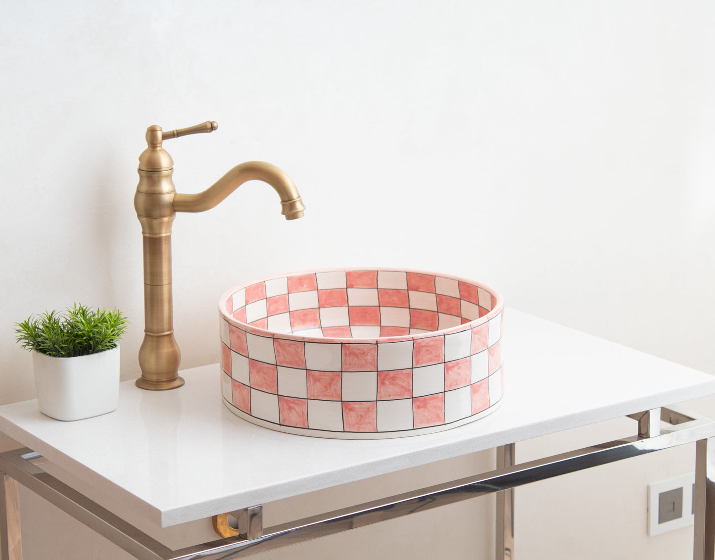 Pink and white checkered ceramic vessel sink on white contemporary vanity with brass traditional faucet and decorative green plant in modern bathroom