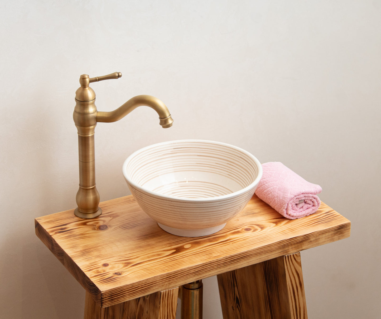Cream ceramic vessel sink with spiral texture styled on rustic wooden vanity with brass faucet and pink towel in bathroom setting