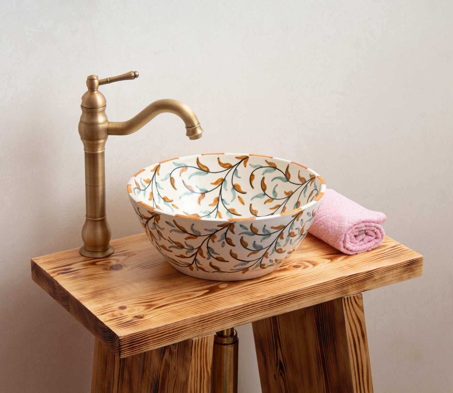 Ceramic vessel sink with hand-painted olive branch design styled on rustic wooden vanity with brass faucet and pink towel