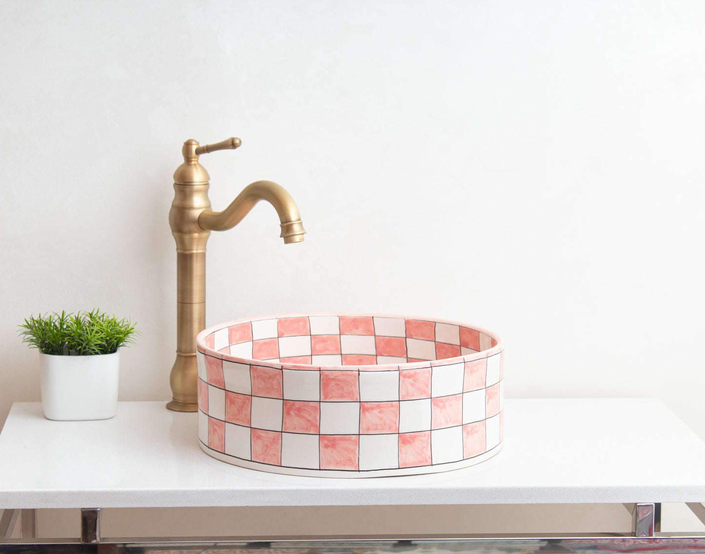 Pink checkered ceramic vessel sink on white floating vanity with tall brass faucet and decorative green plant in contemporary bathroom setting