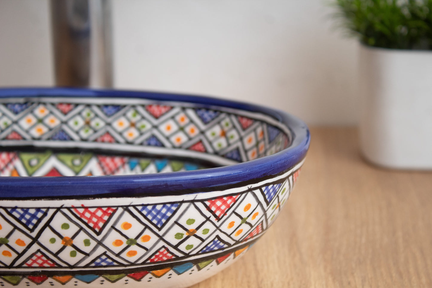 Close-up detail of tiny oval ceramic vessel sink rim highlighting the colorful geometric diamond patterns and blue border on wood surface