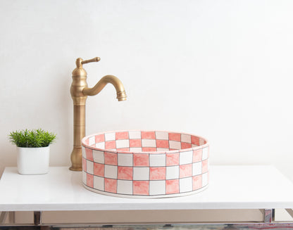 Pink checkered ceramic vessel sink on white floating vanity with tall brass faucet and decorative green plant in contemporary bathroom setting